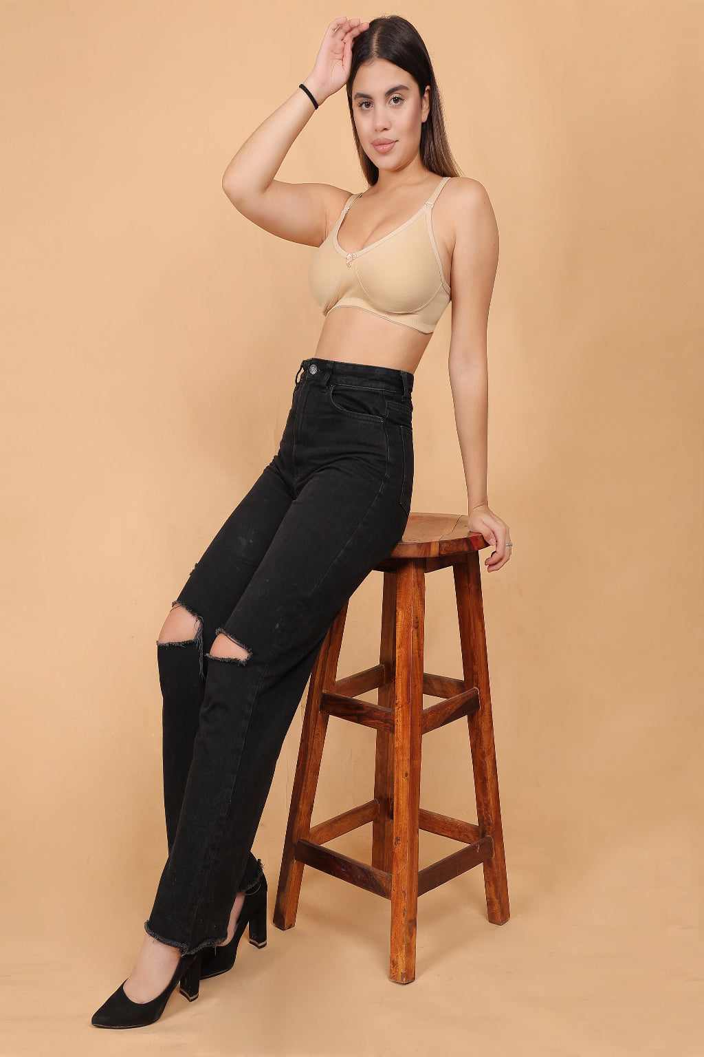 Woman wearing a beige Non padded bra and black ripped jeans sitting on a wooden stool against a beige background